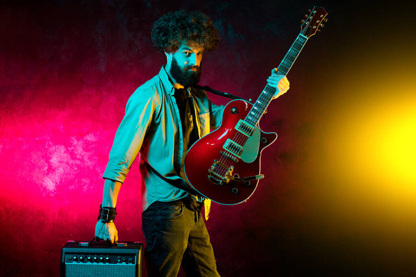 Portrait of hipster man with curly hair with red guitar in neon lights. Rock musician is playing electrical guitar. - Fotó, kép