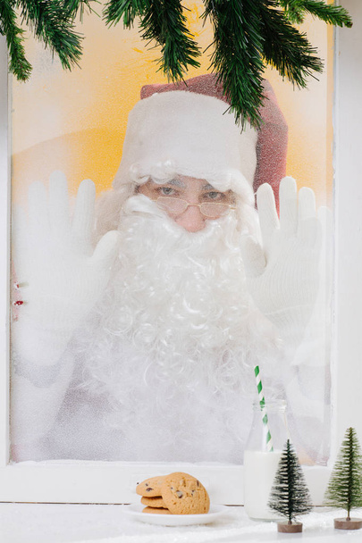 Санта Клаус положил руки в замороженное окно. Вид из дома
. - Фото, изображение