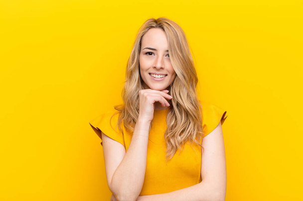 young pretty blonde woman looking happy and smiling with hand on chin, wondering or asking a question, comparing options against flat color wall - Foto, immagini