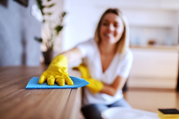 Beautiful young Caucasian blonde housewife with rubber gloves on hands sitting on the floor in living room and dusting shelf. Living room interior. Selective focus on hand. - Photo, Image