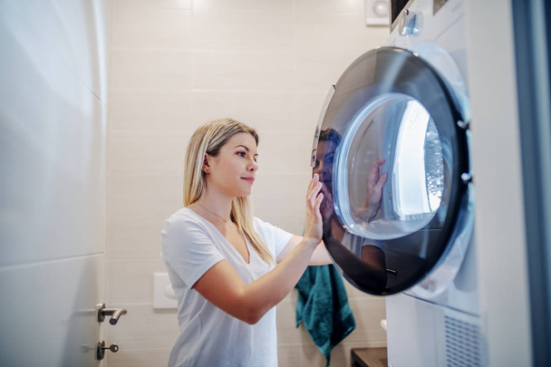 Digne caucasien jeune femme au foyer blonde fermeture machine à sécher tout en se tenant dans la salle de bain dans les dernières heures
. - Photo, image