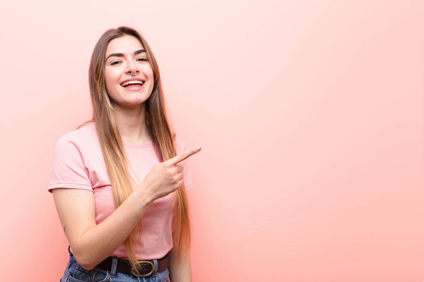 young pretty woman looking excited and surprised pointing to the side and upwards to copy space against pink wall - Photo, Image