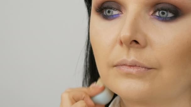 Stylist make-up kunstenaar brengt stichting voor gezicht met een speciale borstel op het gezicht van jonge mooie vrouw met blauwe ogen - Video