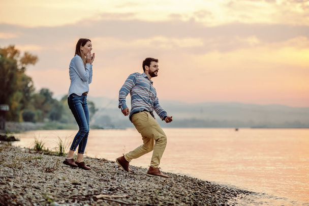 schöner kaukasischer bärtiger Mann, der Steine überspringt, während er an der Küste neben dem Fluss steht. seine reizende Freundin sieht ihm zu und lächelt. - Foto, Bild