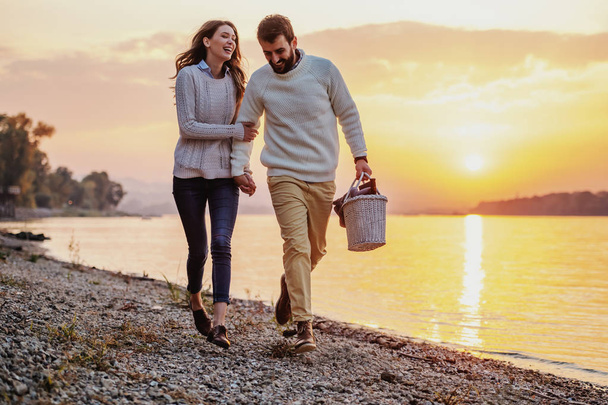 Happy caucasian fashionable couple in love holding hands and walking on coast near river. Man holding picnic basket. In background is sunset. - Zdjęcie, obraz