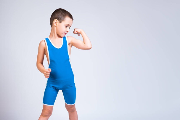 Portrait of a little cheerful boy in a blue  wrestling tights shows biceps, looks confidently at him and poses on a white isolated background. The concept of a little fighter athlete - 写真・画像