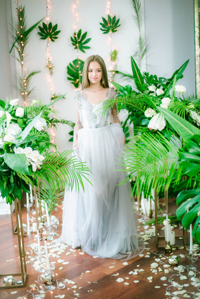 Attractive young girl with blond hair and evening make-up in a romantic dress among tropical greenery and white orchids, roses and lilies. Beauty and fashion - Foto, Imagen