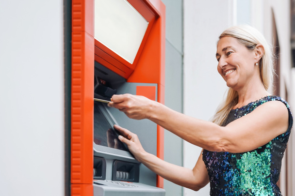 glückliche reife Frau hebt Geld vom Bankautomaten mit EC-Karte ab - Seniorin bezahlt mit Kreditkarte am Geldautomaten - Geschäfts-, Bank- und Lifestylekonzept - Foto, Bild