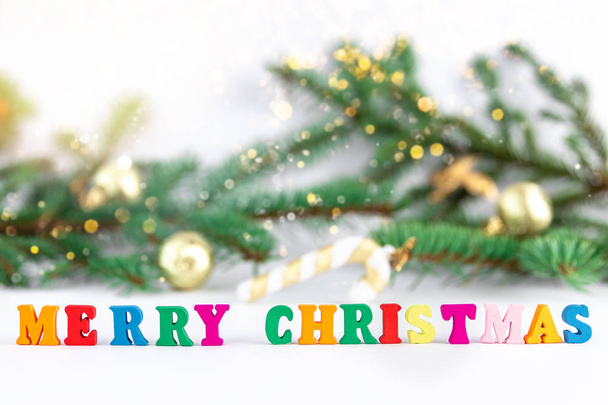 Creative layout of words merry christmas made of wooden multi-colored letters on white background with blurred fir branches, bokeh lights and golden toys, New Year concept, copy space for note or text - Zdjęcie, obraz
