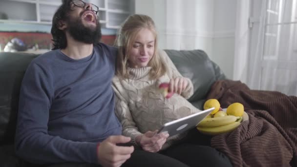 Cheerful married couple looking at tablet screen and laughing out loud. Pregnant Caucasian woman showing her husband something funny and eating apple. Family enjoying time together indoors. - Séquence, vidéo