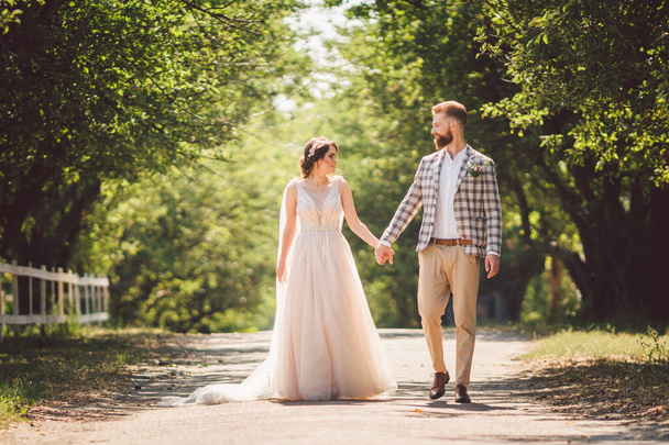 Małżeństwo weselne lubi spacerować po lesie. Nowożeńcy przytulają się i trzymają za ręce. Nowożeńcy idą do przodu trzymając się za rękę w parku wzdłuż ścieżki w słoneczną pogodę. Temat ślubu i zaręczyn - Zdjęcie, obraz