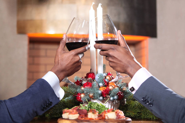 Duas mãos afro-americanas com copos de vinho tinto em foco. Mãos levantadas no fundo da composição de Natal com velas. O conceito de reuniões de negócios. Ano Novo
. - Foto, Imagem