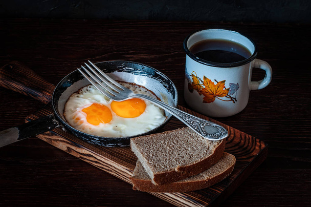 Fried scrambled eggs in vintage pan, with bread, old fork, metal mug with tea, top view - Photo, Image