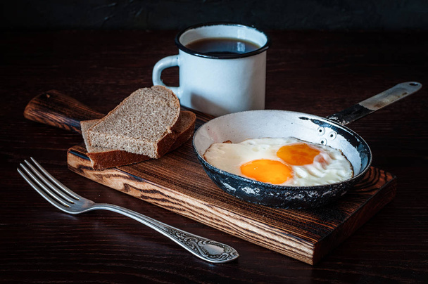 Fried scrambled eggs in vintage pan, with bread, old fork, metal mug with tea, side view, dark background - Photo, Image