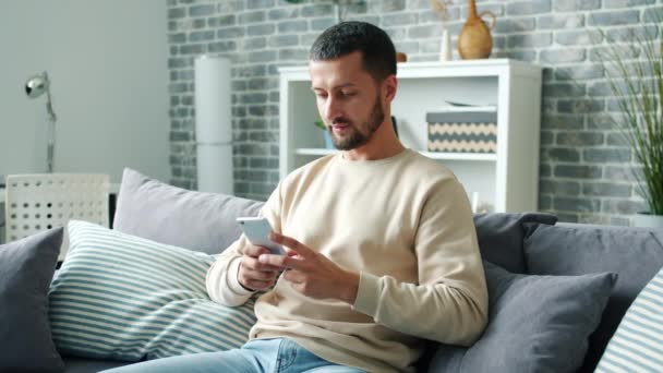 Joyful bearded man using smartphone texting having fun smiling in house alone - Imágenes, Vídeo