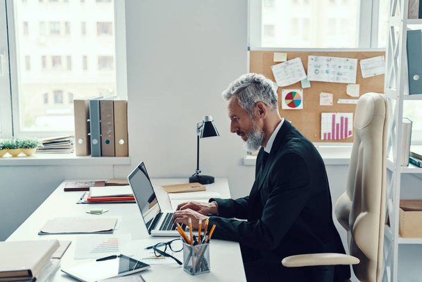 Концентрований зрілий чоловік у повному костюмі використовує ноутбук під час роботи в сучасному офісі
 - Фото, зображення