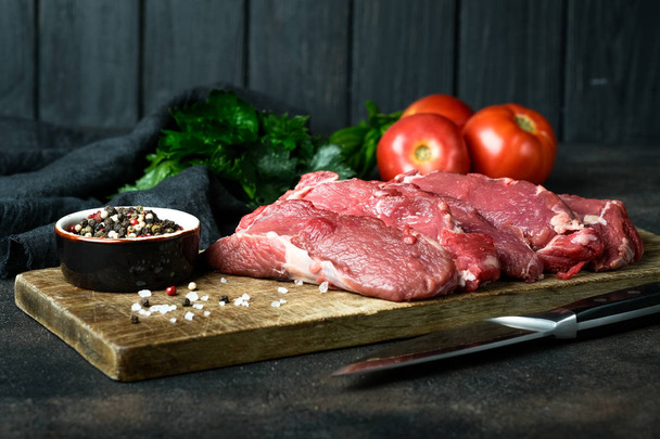 Carne fresca cruda, tagliata in bistecche con verdure, erbe aromatiche e spezie su una tavola di legno su fondo scuro
.  - Foto, immagini