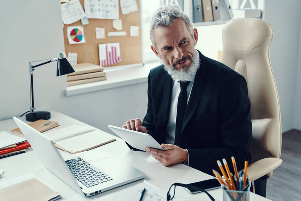 Thoughtful mature man in full suit using digital tablet and looking away while working in modern office - Photo, image