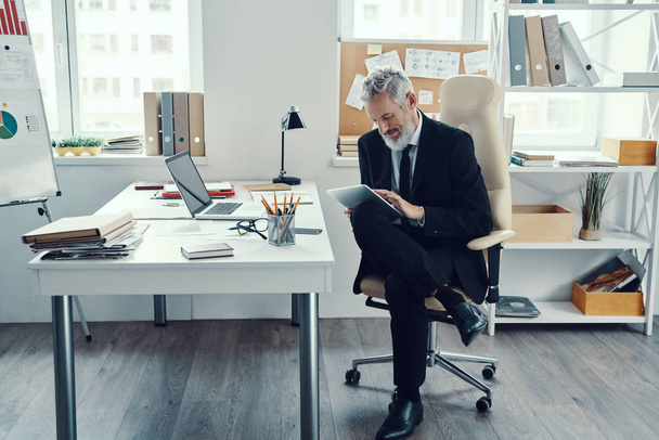 Thoughtful mature man in full suit using digital tablet while working in modern office - Foto, Bild