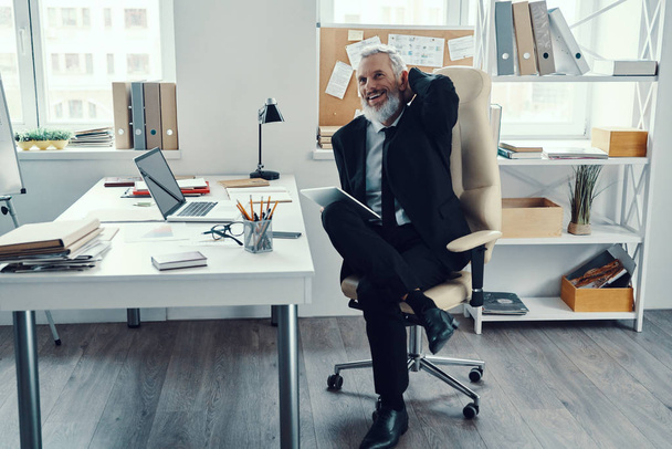 Heureux homme mature en costume complet à l'aide d'une tablette numérique et souriant tout en travaillant dans un bureau moderne
 - Photo, image