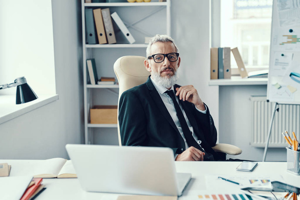 Confident mature man in full suit looking at camera while working in modern office - Foto, Bild
