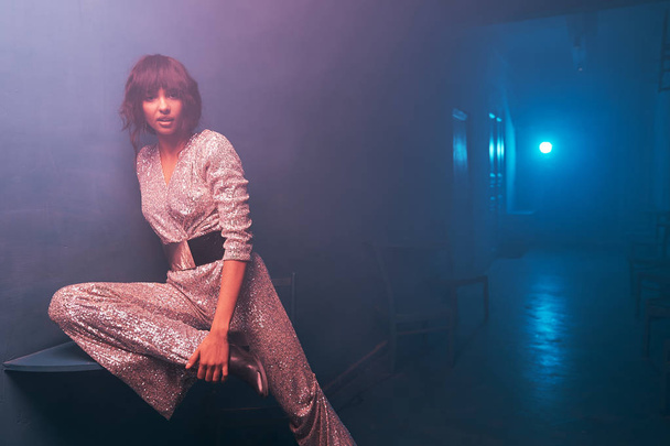 dark-haired girl sitting on a table in blue and violet light in a silver jumpsuit with a wide belt and cowboy shoes, she has silver big earrings, professional makeup and looks into the camera - Foto, Bild