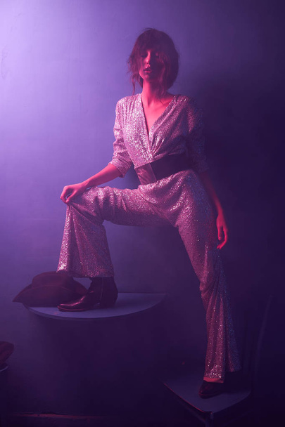 dark-haired girl stands in a blue light on chair in a silver jumpsuit with a wide belt, she has cowboy shoes and a hat on the table - Foto, immagini