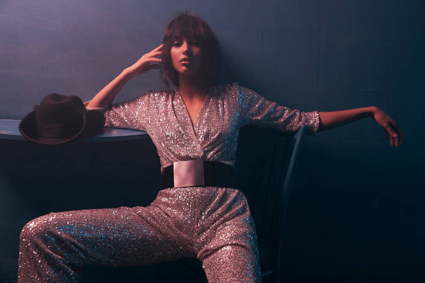 dark-haired girl sitting on a chair in violet light in a silver jumpsuit with a wide belt, she has professional makeup and looks in camera - Foto, afbeelding