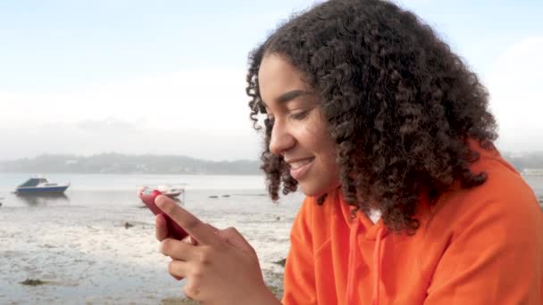 Beautiful mixed race African American girl teenager young woman wearing orange hoodie, sitting on a sea front using her smart cell phone for social media or text messaging - Πλάνα, βίντεο