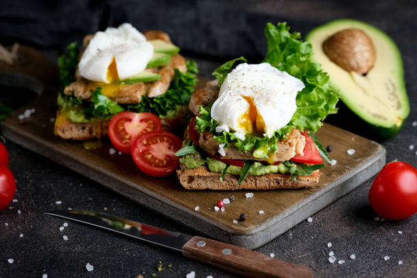 アボカドとポーチドエッグのおいしいサンドイッチ,濃い背景に緑の葉とトマトと.健康的な朝食.  - 写真・画像