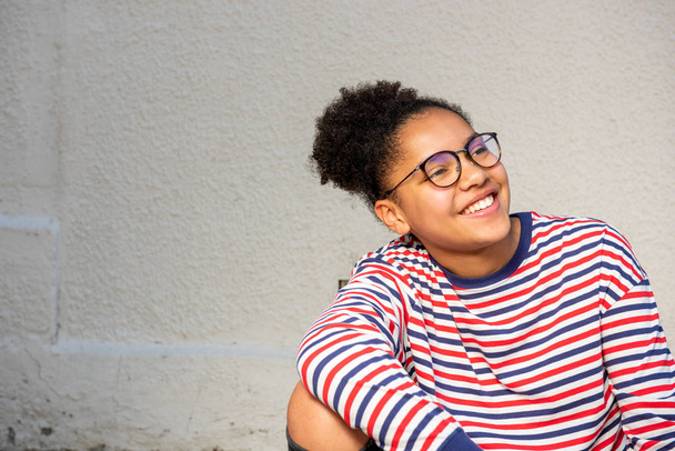 Gözlüklü, beyaz duvarda oturan gülümseyen Afrikalı Amerikalı genç kızın yakın portresi. - Fotoğraf, Görsel