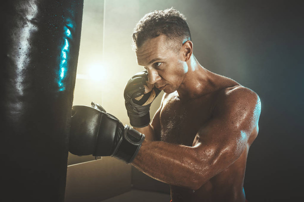 Keskittynyt lihaksikas mies harjoittelee aktiivisesti ja kovaa, lyömällä nyrkkeilysäkki
. - Valokuva, kuva