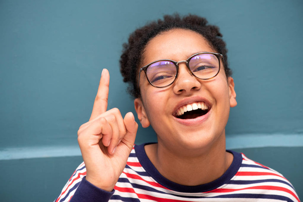 Close up portrait of smiling young mixed race girl with glasses pointing finger up against blue wall - Photo, Image