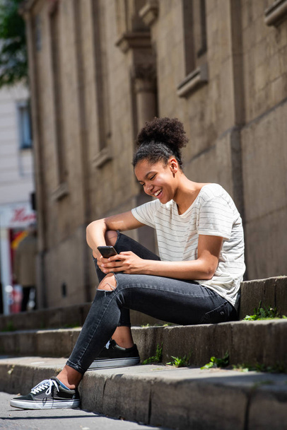 Portret van een glimlachend jong Afrikaans Amerikaans meisje dat buiten zit en naar een mobiele telefoon kijkt - Foto, afbeelding