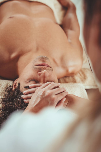 Bonito homem está deitado na mesa de massagem e recebendo massagem facial saudável por terapeuta jovem no centro de spa
. - Foto, Imagem
