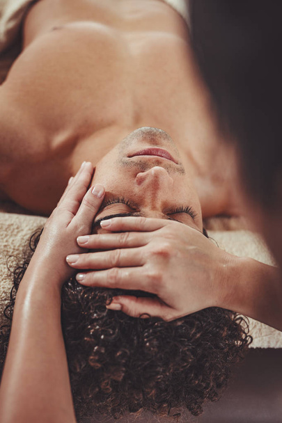 Handsome man is lying down on massage table and getting healthy facial massage by young female therapist in the spa center. - Fotografie, Obrázek