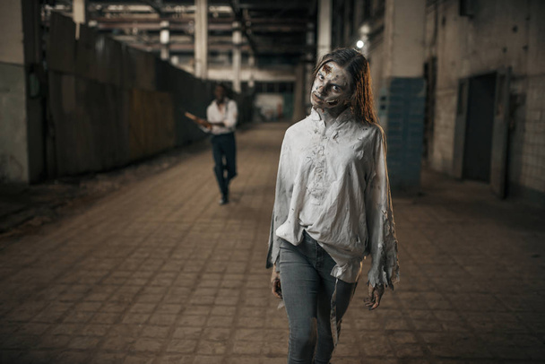 Man with axe catching up female zombie in abandoned factory, scary place. Horror in city, creepy crawlies, doomsday apocalypse, bloody evil monsters - Photo, Image