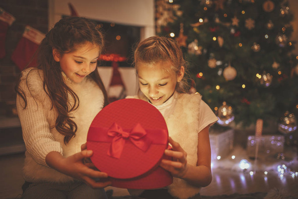 Zwei lächelnde Mädchen sitzen auf dem Boden neben dem Weihnachtsbaum. Überrascht betrachten sie das Geschenk in einer roten Schachtel - Foto, Bild