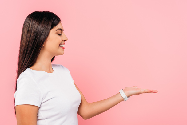 side view of smiling pretty brunette girl pointing with hand at copy space isolated on pink - Foto, Imagem