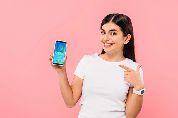 lächelndes hübsches brünettes Mädchen zeigt mit Finger auf Smartphone mit Online-Shopping-App isoliert auf rosa - Foto, Bild