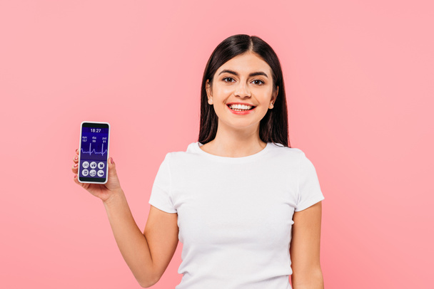  smiling pretty brunette girl holding smartphone with healthcare app isolated on pink - Fotoğraf, Görsel