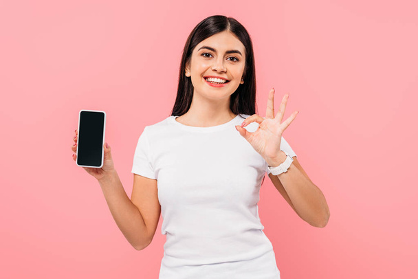 smiling pretty brunette girl holding smartphone with blank screen and showing ok isolated on pink - Foto, Bild