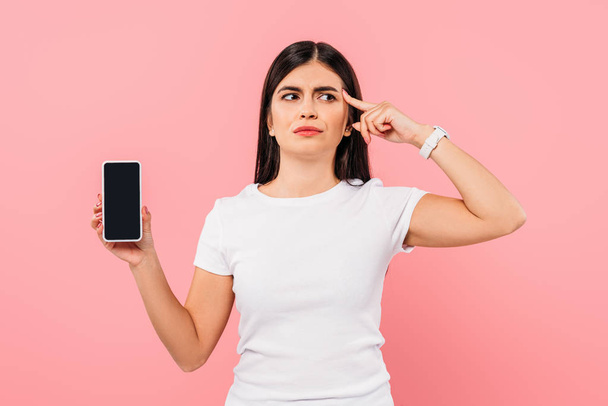 thoughtful pretty brunette girl holding smartphone with blank screen isolated on pink - Φωτογραφία, εικόνα