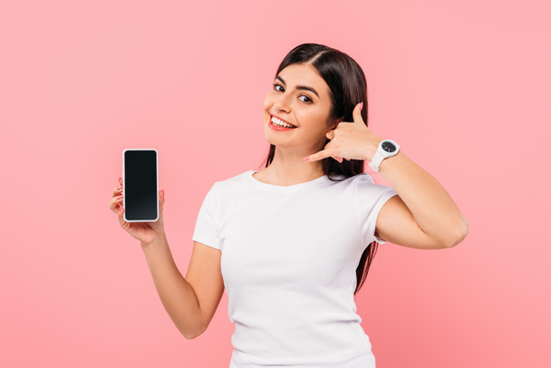 sonriente chica morena bonita sosteniendo teléfono inteligente con pantalla en blanco y mostrando gesto de llamada aislado en rosa
 - Foto, Imagen