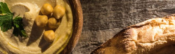 vista superior de delicioso hummus com grão de bico em tigela perto de pita cozido na mesa rústica de madeira, tiro panorâmico
 - Foto, Imagem