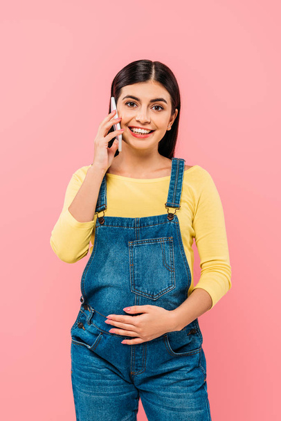 smiling pregnant pretty girl talking on smartphone with hand on belly isolated on pink - Photo, Image