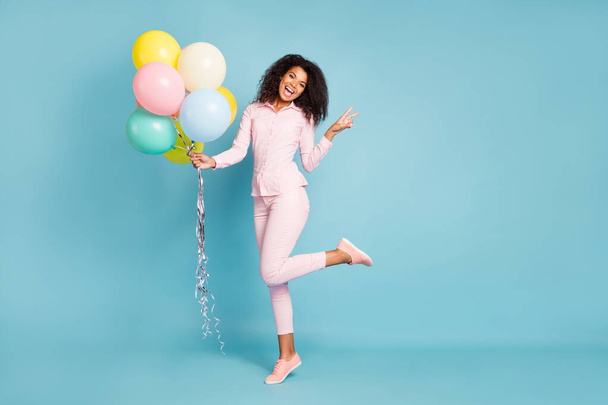 Full length photo of amazing wavy dark skin model lady holding many air balloons showing v-sign symbol good mood wear pink shirt trousers isolated blue color background - Fotoğraf, Görsel
