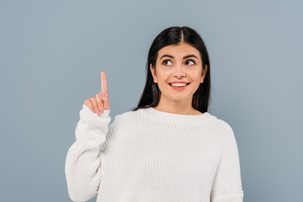 smiling pretty brunette girl in white sweater pointing with finger upwards isolated on grey - Foto, Imagem