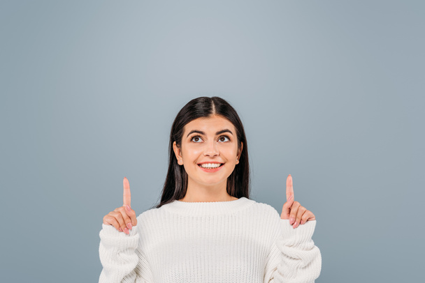 smiling pretty brunette girl in white sweater pointing with fingers upwards isolated on grey - Foto, Imagem