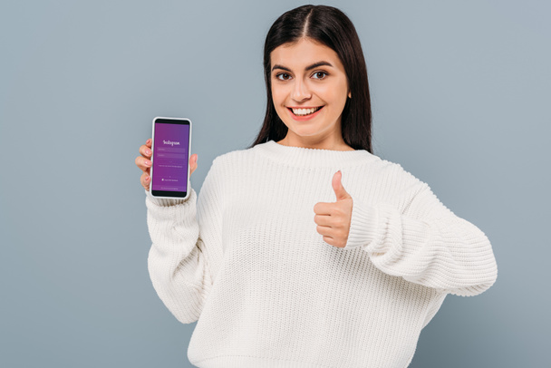 usmívající se hezká dívka v bílém svetru ukazuje smartphone s instagram app a palec nahoru izolované na šedé - Fotografie, Obrázek